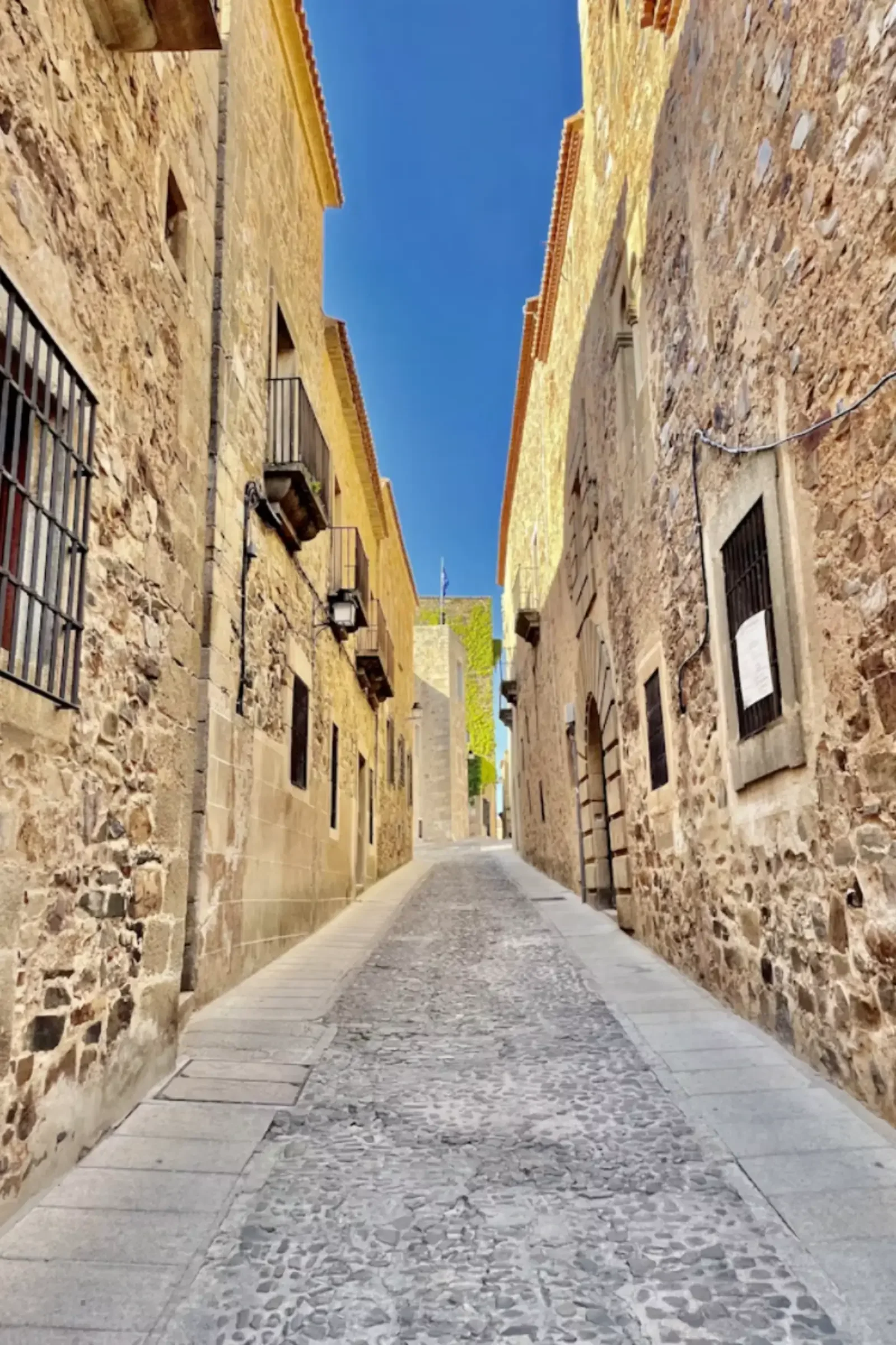 A narrow cobblestone street flanked by old stone buildings with small windows and wrought iron details. The sky is clear and blue at the end of the street.
