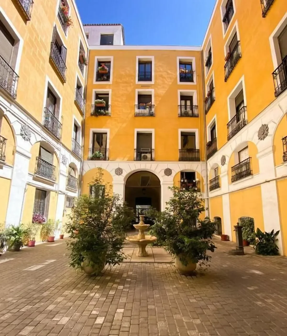 Courtyard with potted plants, a fountain, and a yellow building with multiple windows and balconies under a clear blue sky.