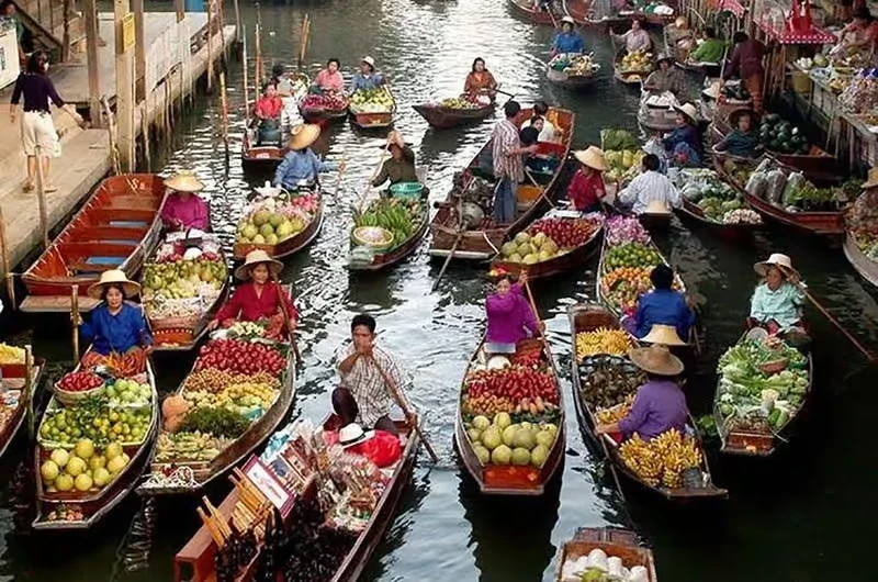 Market boats filled with fresh fruits and vegetables on a canal, with vendors and shoppers wearing traditional clothing and hats.