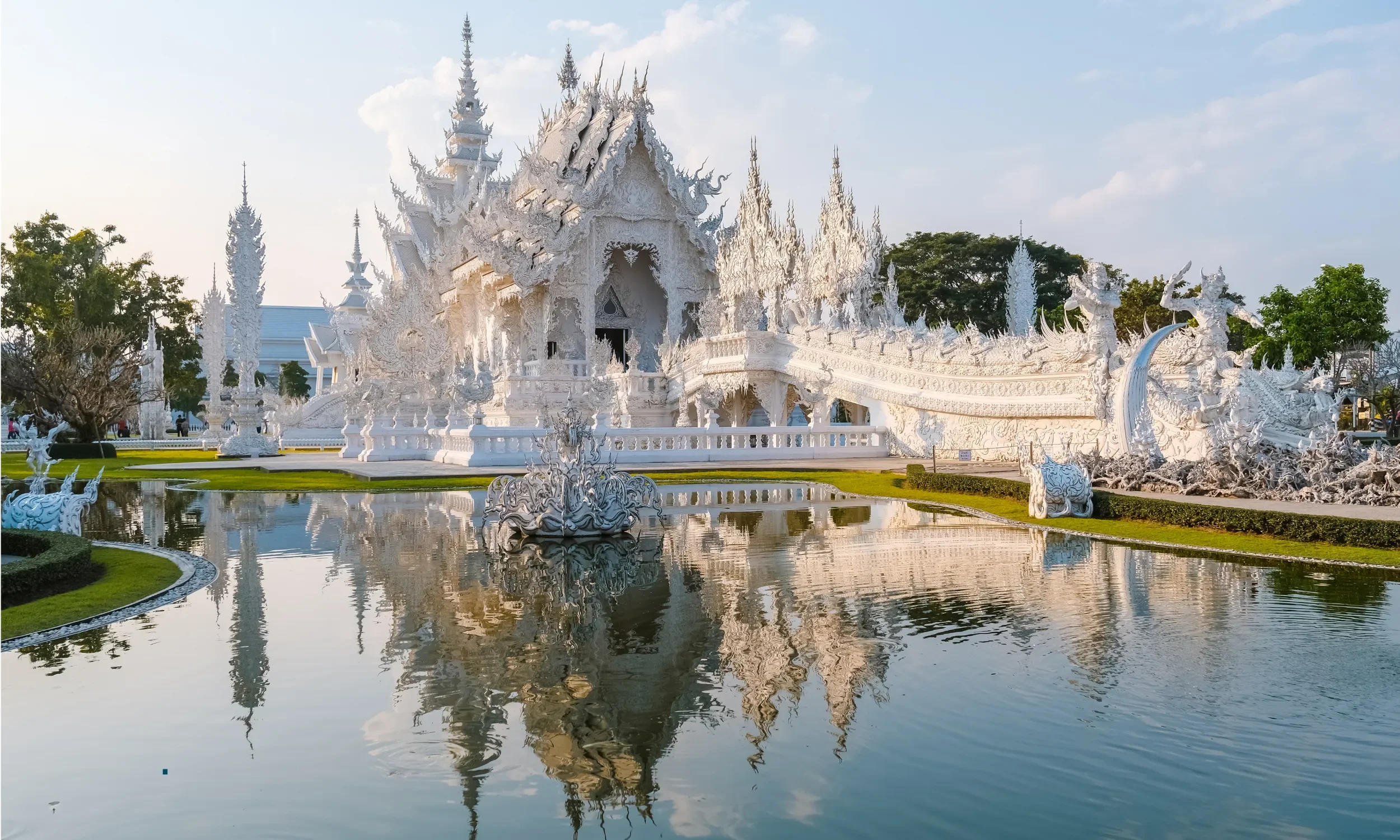 An ornate white temple with intricate details, surrounded by a pond that reflects the structure, set against a background of trees and a partly cloudy sky.