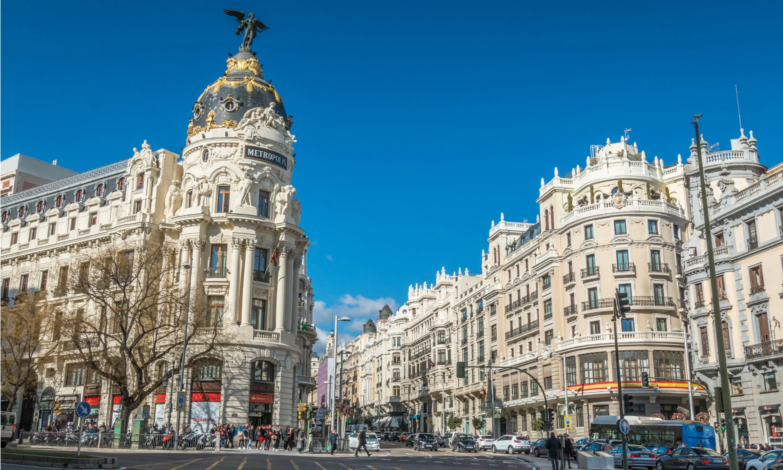 Street view of ornate white buildings with balconies and decorative sculptures, with a clear blue sky above and a busy crosswalk with pedestrians and cars.