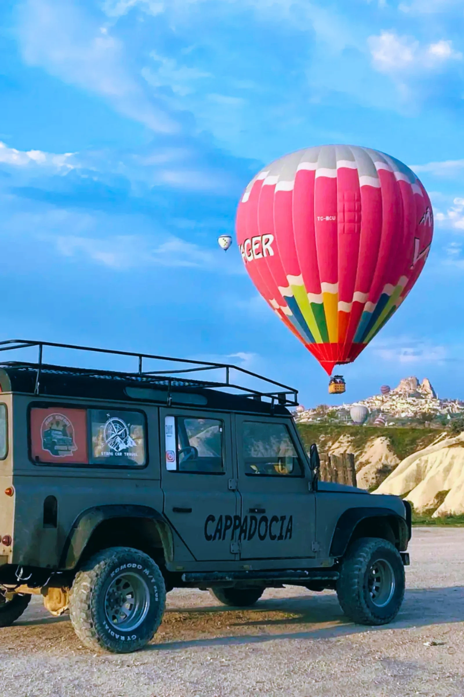 A hot air balloon with pink, white, and gray colors flying in a blue sky with scattered clouds. A smaller hot air balloon is visible further away. In the foreground, a black off-road vehicle with the word 'Cappadocia' on its side is parked on a dirt area, with rocky formations and buildings in the background.