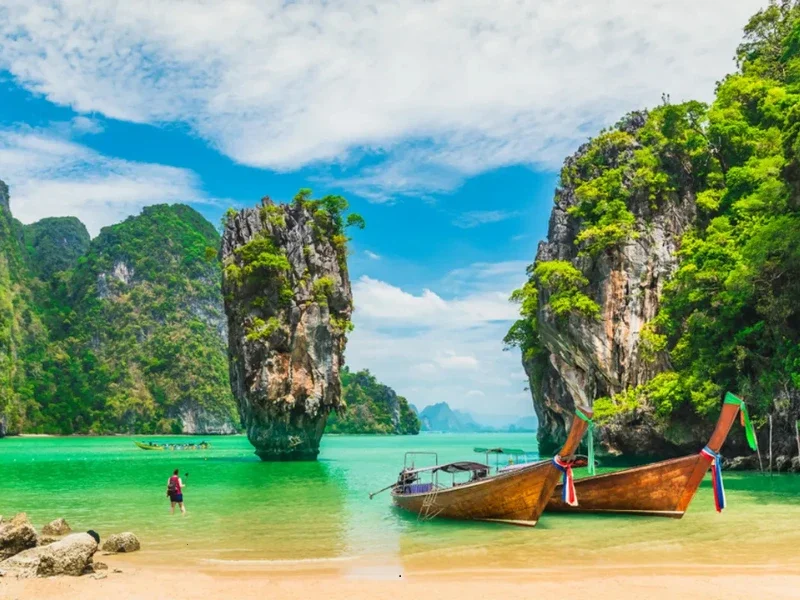 Tropical beach with clear turquoise water, large limestone cliffs covered in greenery, a traditional wooden boat decorated with colorful ribbons, and a person in swimwear near the water, under a partly cloudy sky.