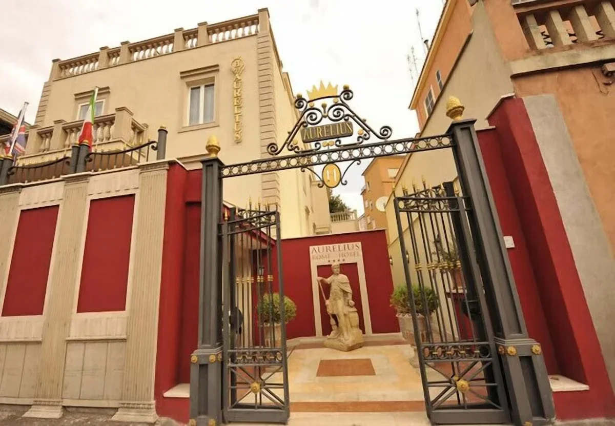 Entrance gate to Aurelius Roman Hotel with a statue of a Roman soldier behind the gate, surrounded by potted plants and Roman-style decorative elements.