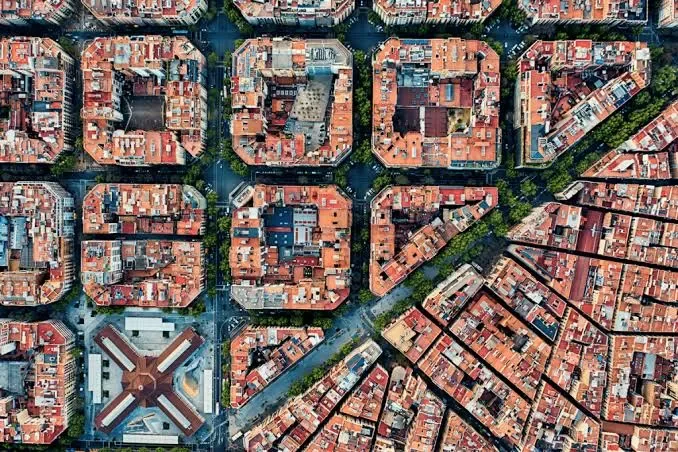 Aerial view of a dense urban city block with red-tiled roofs, grid-like streets, and a large building with a cross-shaped roof at the intersection.