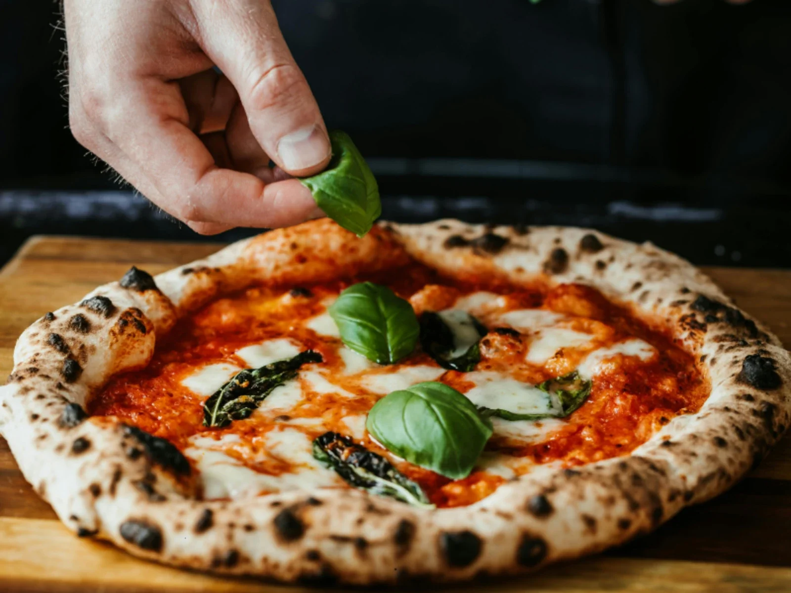 A hand sprinkling fresh basil leaves onto a cooked pizza with cheese, tomato sauce, and basil leaves, on a wooden surface.