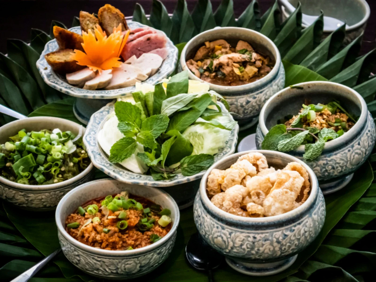 A traditional Thai meal with multiple small bowls containing various sauces, herbs, and condiments, arranged on a dark table with a large green leaf underneath.