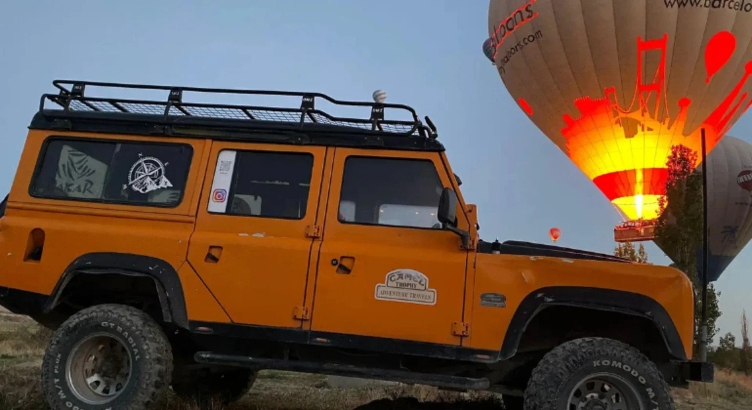 An orange off-road vehicle with a roof rack parked on a dirt terrain, with hot air balloons illuminated and floating in the sky in the background.
