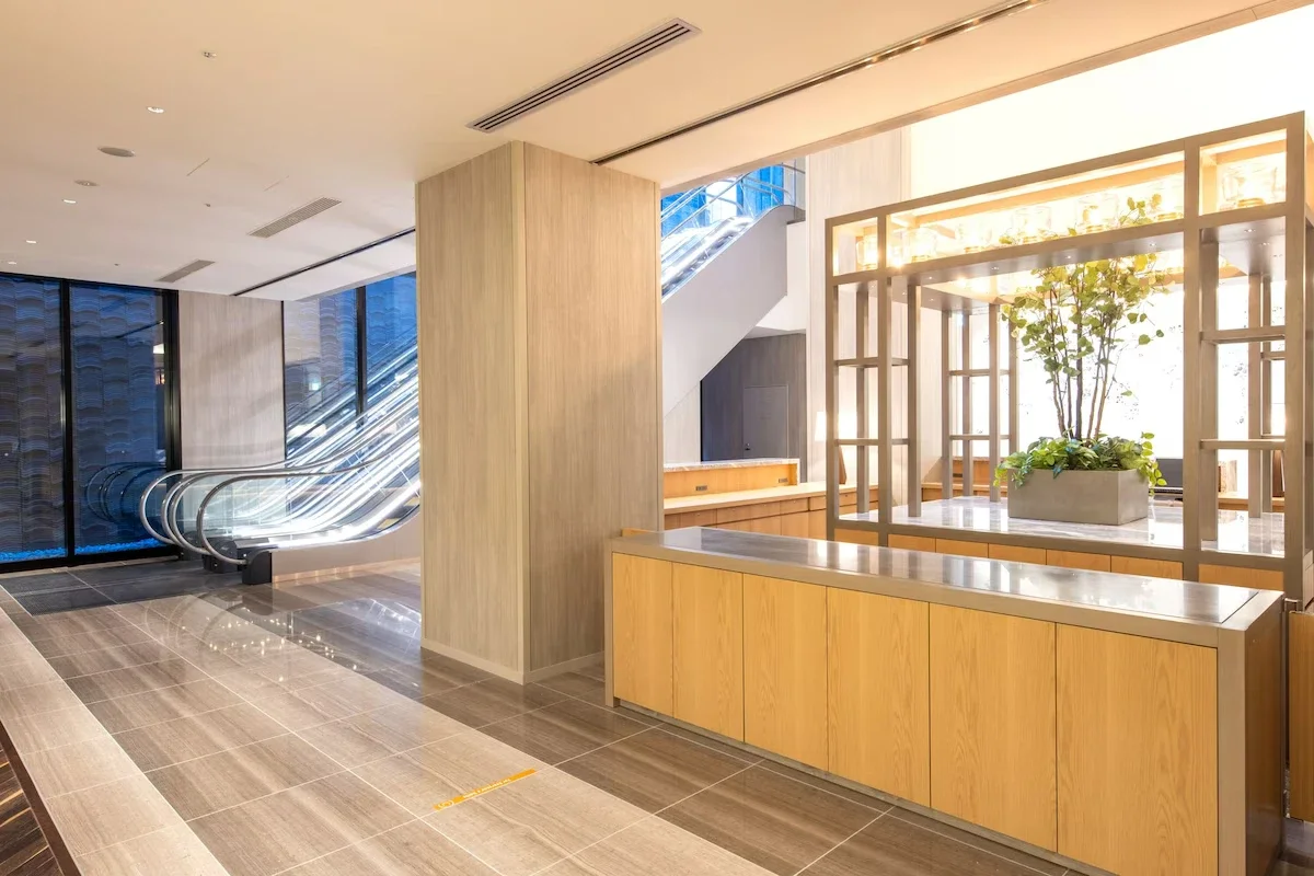 Interior view of a modern building lobby with a staircase, escalators, large windows, and a decorative planter with greenery.