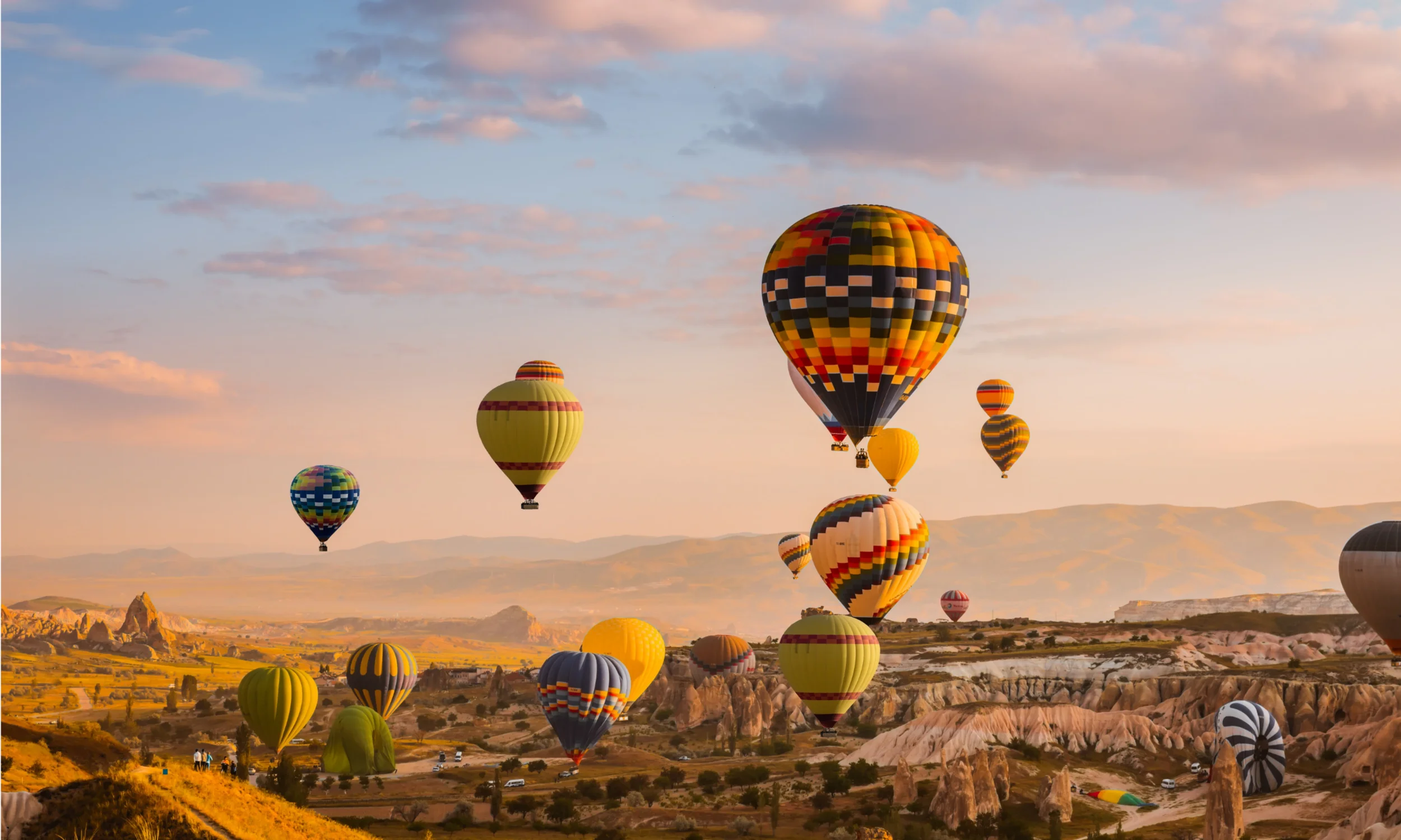 Multiple colorful hot air balloons floating over a valley at sunrise with scattered clouds in sky.