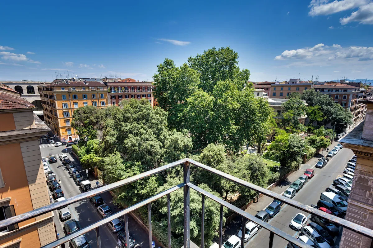 An urban view from a balcony showing a street lined with parked cars, several large trees, and buildings under a partly cloudy blue sky.