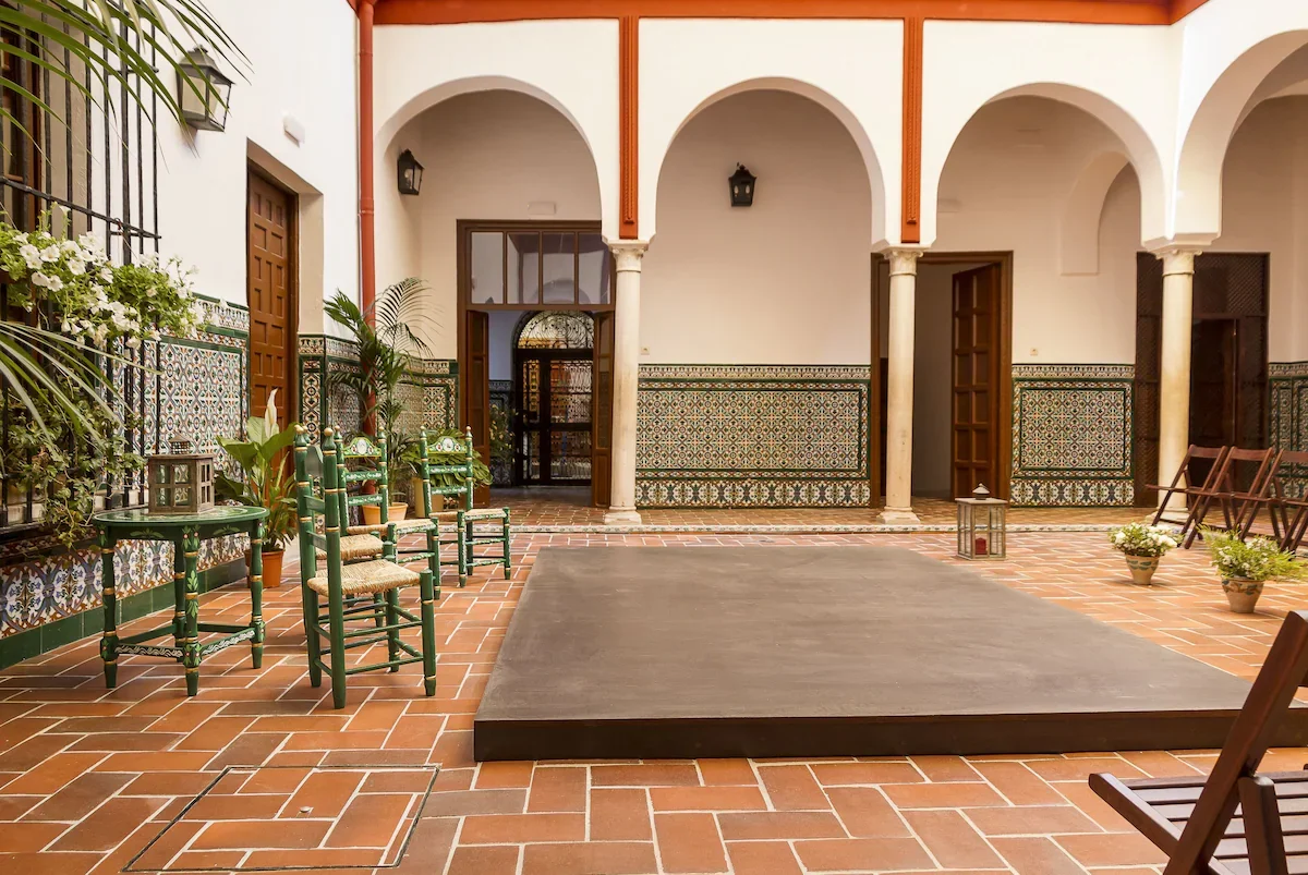 Indoor courtyard with terracotta tile flooring, decorated with potted plants, green wooden chairs, a small table, and a stage, featuring arched doorways and tiled walls with intricate patterns.
