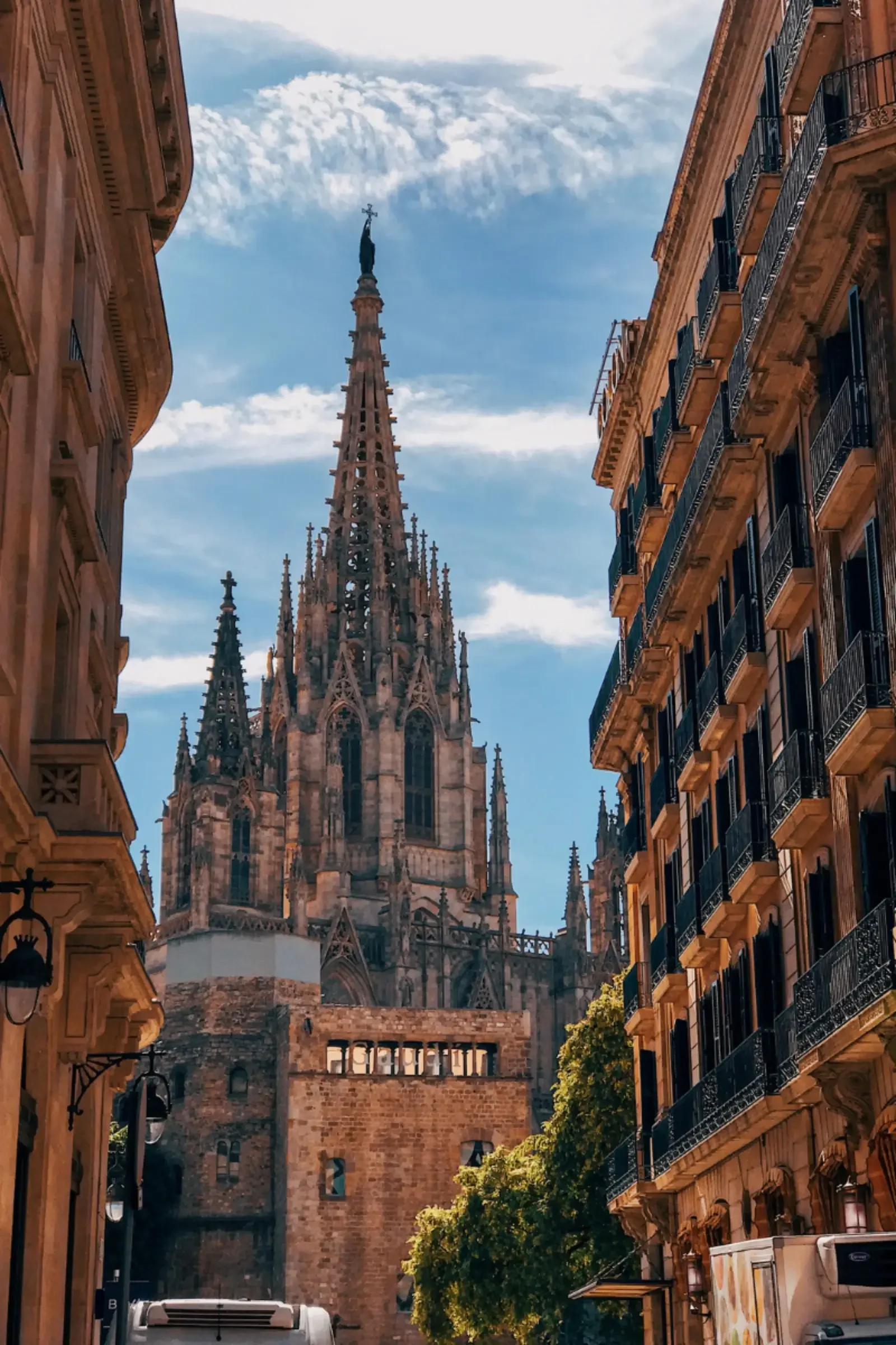A narrow street in Barcelona with classic European buildings on both sides, featuring wrought-iron balconies, and a Gothic cathedral with a tall spire in the background under a partly cloudy sky.