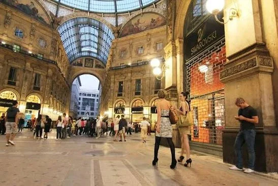 Indoor shopping mall with high arched glass ceiling, multiple store entrances, and people walking and shopping.