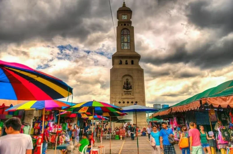 A vibrant outdoor market with colorful umbrellas, many vendors selling goods, and a tall clock tower under a cloudy sky.