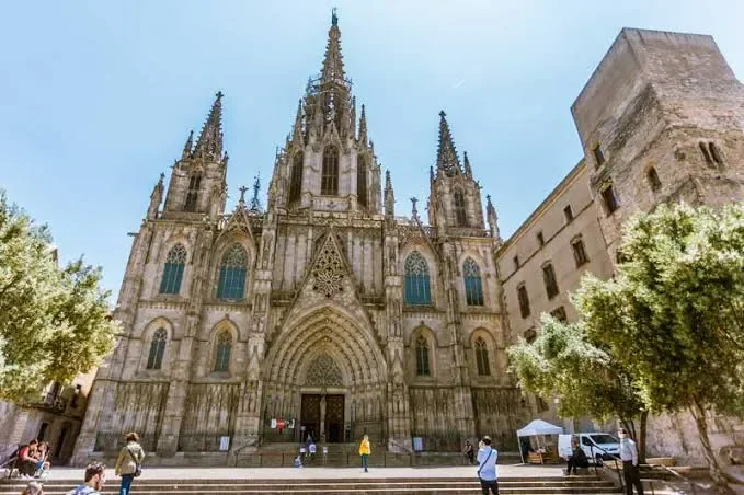A large Gothic cathedral with intricate stone details and tall spires, situated on a city street with trees and pedestrians.