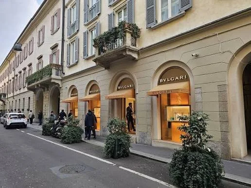 Street scene with luxury boutiques Bvlgari, pedestrians, parked cars, and potted plants outside a building with tall windows and balconies.