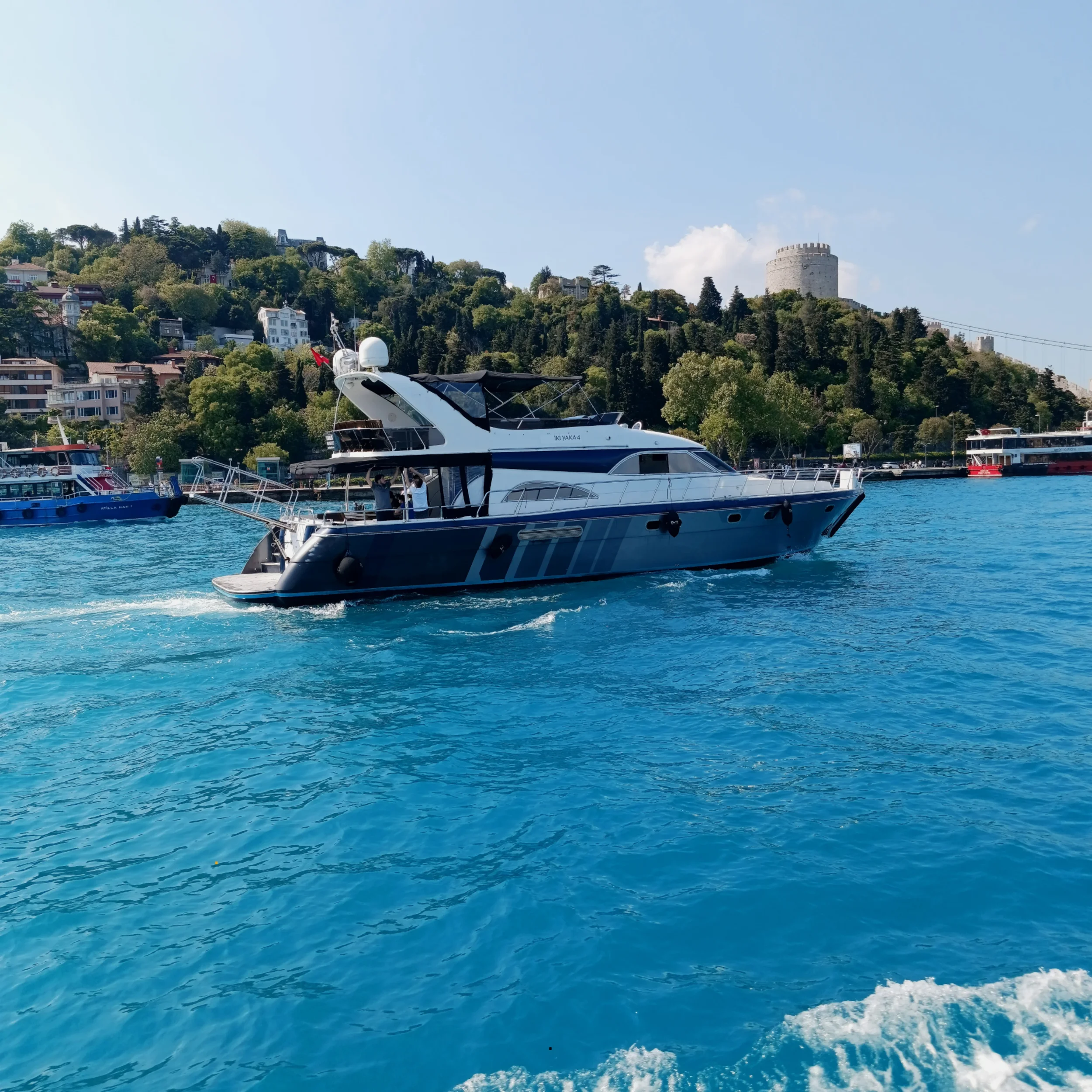 Motor yacht cruising on blue water with hillside buildings and a castle in the background under a partly cloudy sky.