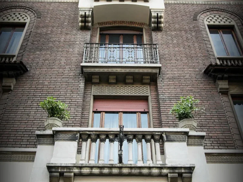 Close-up of a brick building's facade with decorative architectural elements, small balconies, potted plants, and windows.