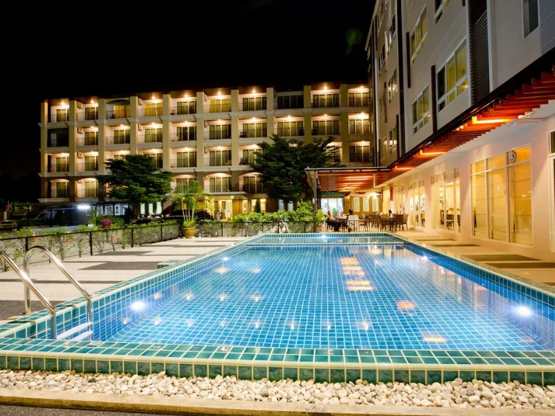 Night view of a hotel with a lit outdoor swimming pool, surrounded by deck chairs, tables, and chairs, with a multi-story building in the background and trees.