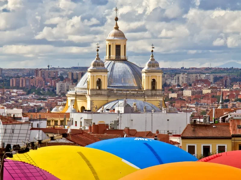 Colorful umbrellas in the foreground with a historic cathedral featuring a large silver dome and three smaller spires in the background, cityscape with buildings and hills under a cloudy sky.