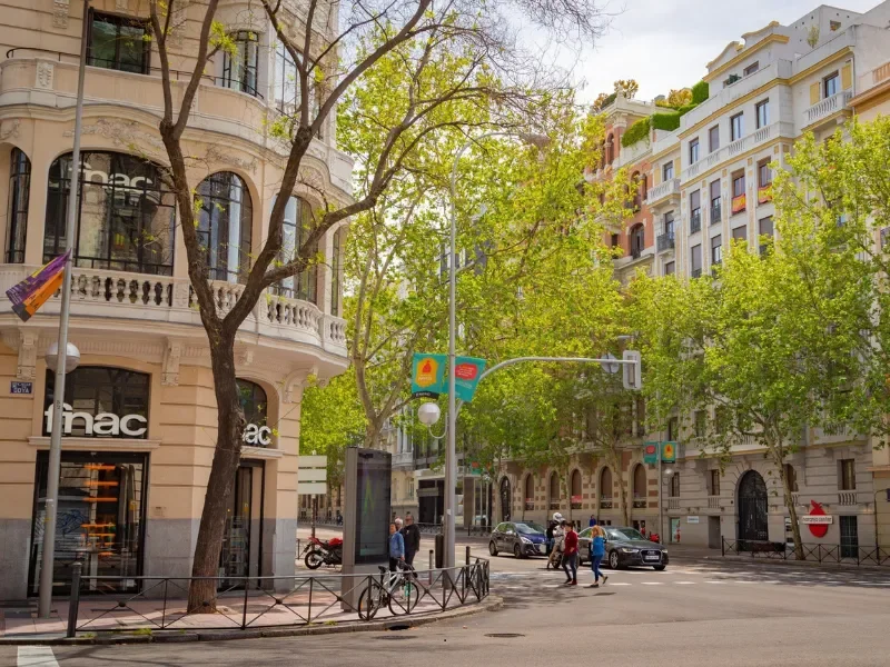 Street scene in an urban city with pedestrians, vehicles, and trees lining the sidewalk, with classic European-style buildings in the background.