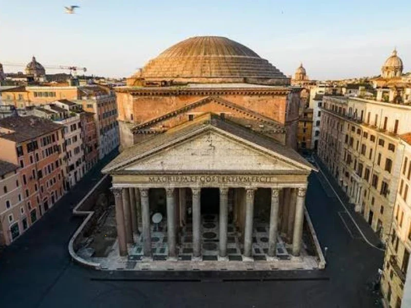 A historic ancient Roman or Greek-style temple with tall columns and a pediment, situated in an urban setting with surrounding buildings and a large domed structure behind it.