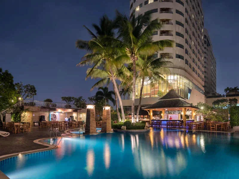 Nighttime view of a hotel pool area with palm trees, a thatched-roof cabana, and a high-rise hotel building illuminated in the background.