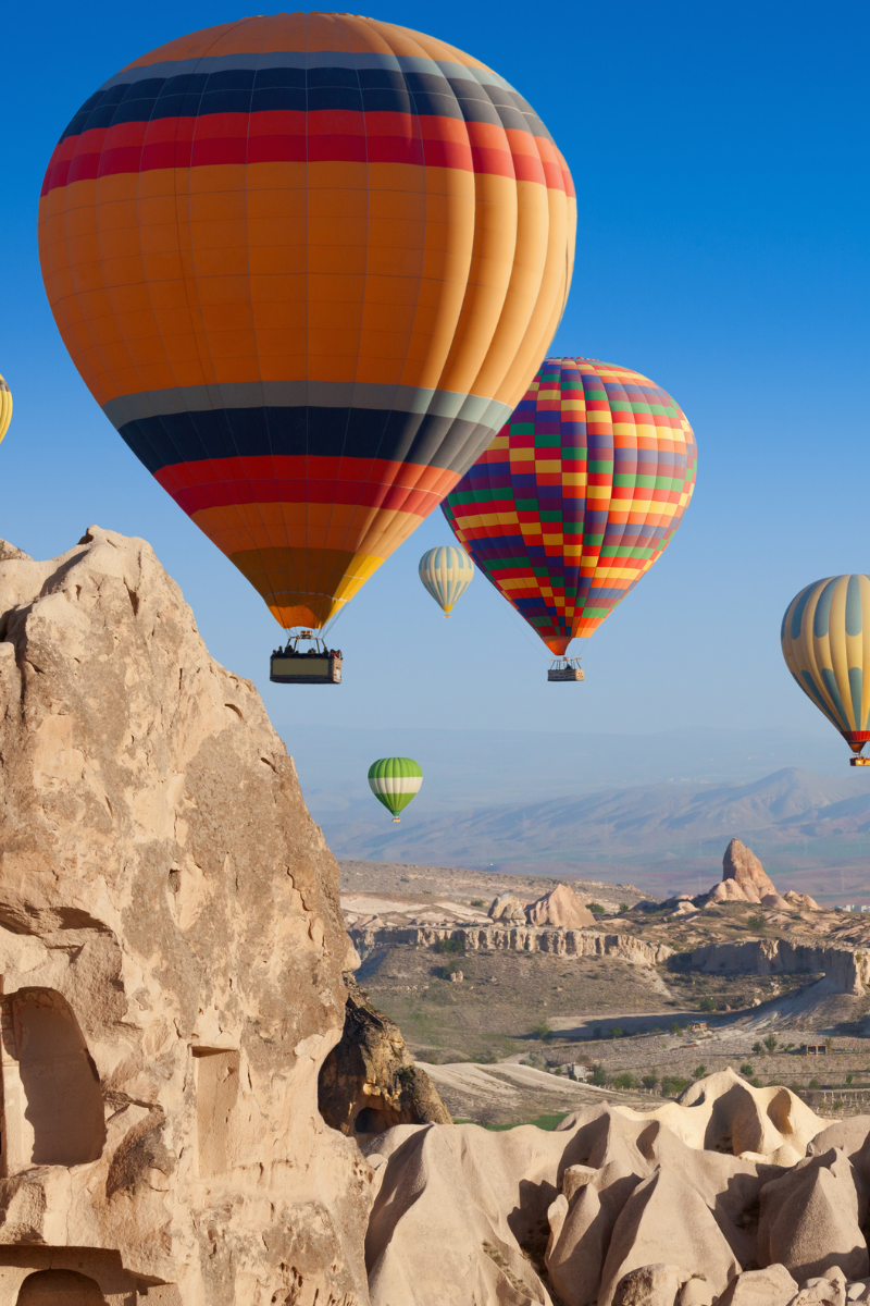 Multiple colorful hot air balloons flying over rocky landscape and rock formations under a clear blue sky.