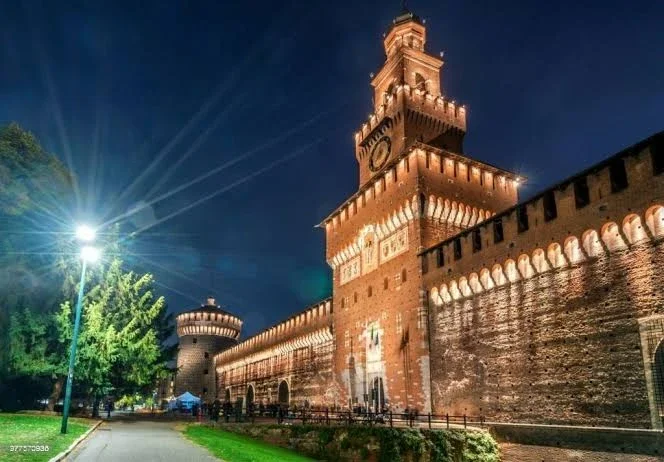 Nighttime view of a historic brick castle with a tall clock tower and battlements, illuminated by exterior lights, with a stone wall extending along the right side and a park with a streetlamp and trees in the foreground.