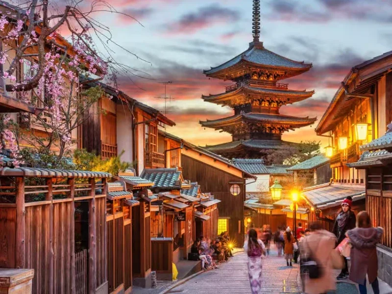Traditional Japanese street with wooden buildings and a pagoda, during sunset with people walking and wearing kimonos.