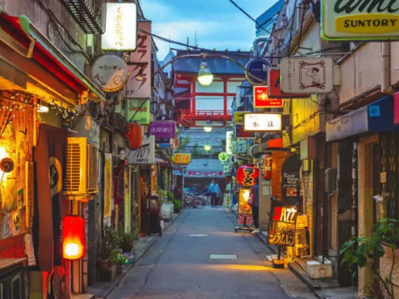 A narrow street in an Asian city, lined with colorful lit storefronts, signs in Japanese, lanterns, and a red torii gate in the distance at dusk.