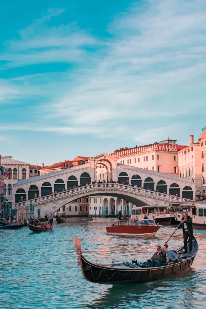 Venetian canal scene with gondolas, pedestrian bridge, and historic buildings under a blue sky.