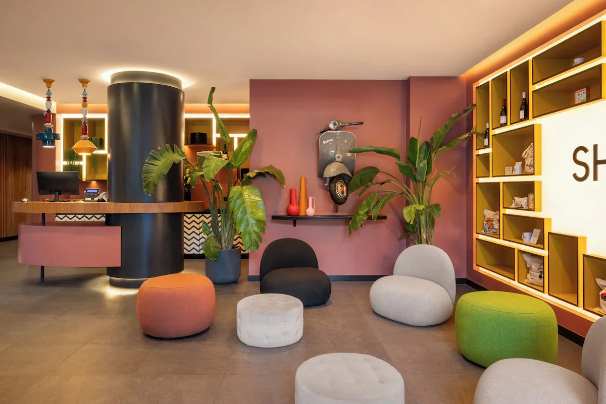 Modern hotel lobby with pink walls, colorful seating, large green plants, and decorative shelving.