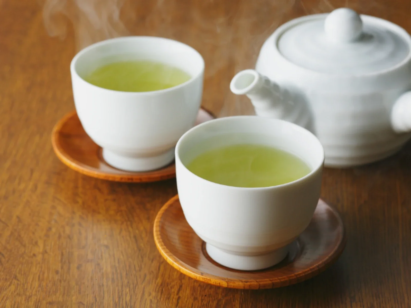 Two white teacups filled with green tea on round wooden saucers, placed on a wooden table with a white teapot in the background.