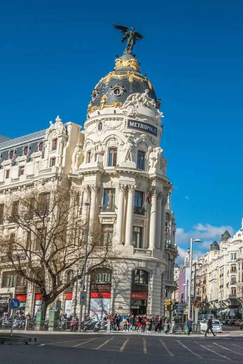 A historic ornate building with a statue on top, labeled 'Metropolis,' with a crowd of people walking past on a city street under a clear blue sky.