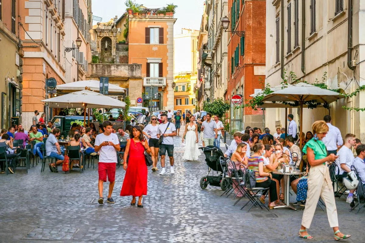 People dining at outdoor cafes on a cobblestone street in a European city, with buildings and colorful facades in the background.