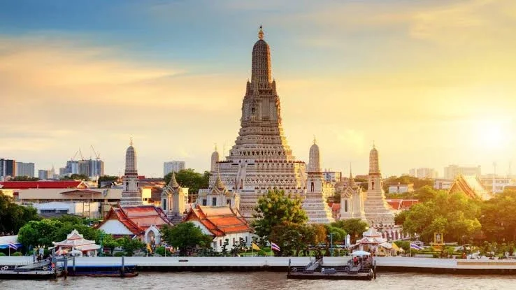 Sunset over Wat Arun, a historic Buddhist temple in Bangkok, Thailand, seen across the Chao Phraya River with trees and city buildings in the background.