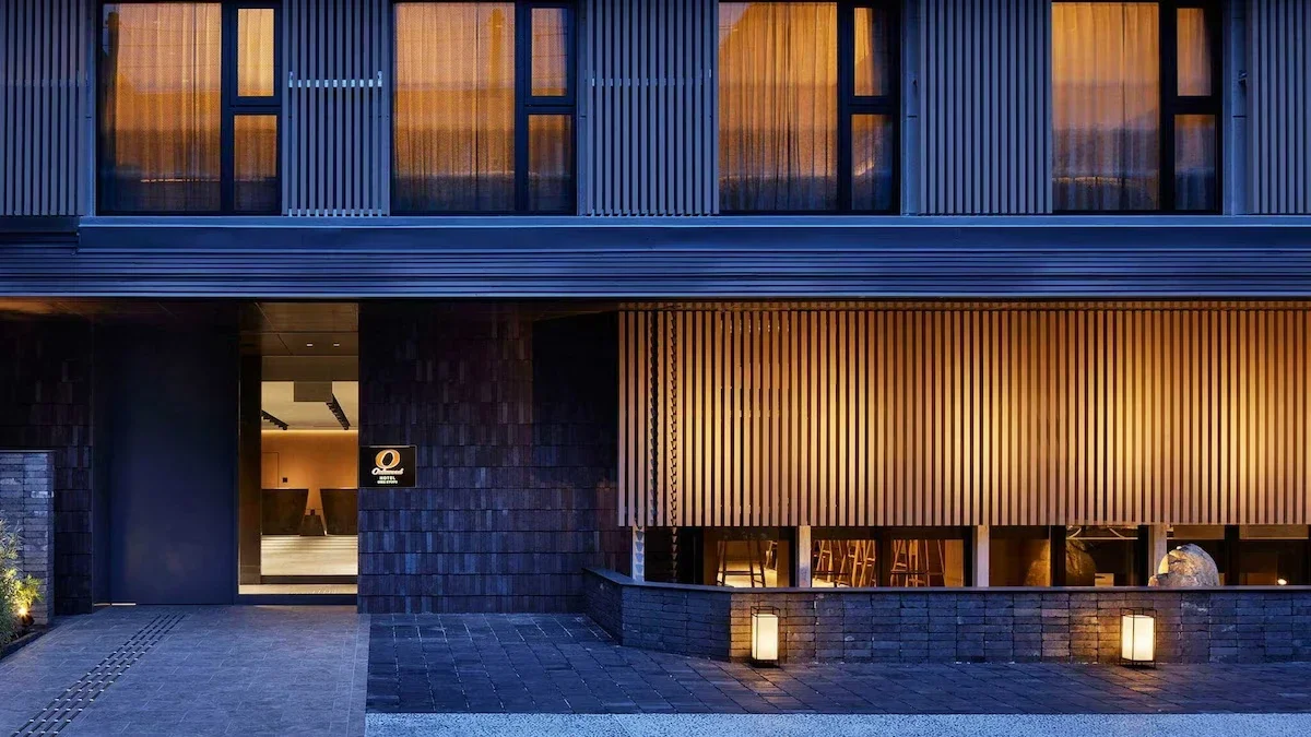 Modern building entrance with dark brick wall and wooden paneling, illuminated by soft outdoor lighting at dusk.