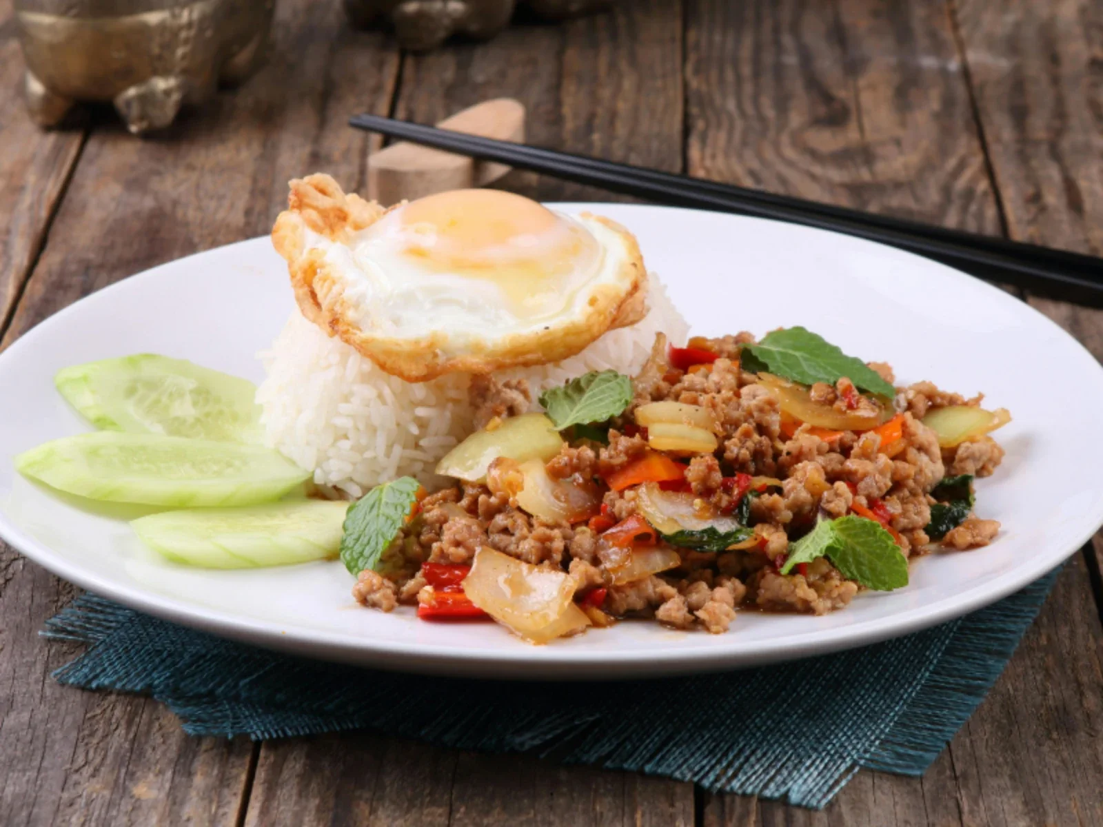A plate of Thai basil ground chicken with rice, a fried egg on top, and cucumber slices on the side, on a wooden table.