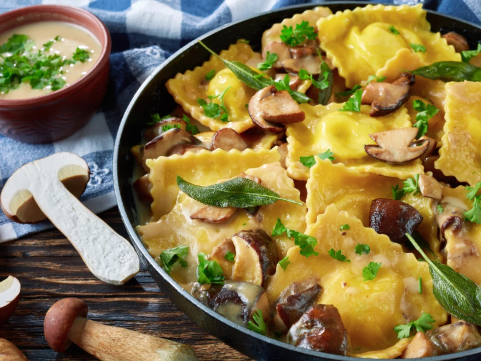 A pan of cooked ravioli garnished with fresh herbs, sliced mushrooms, and leafy greens. A small bowl of creamy sauce or soup with chopped herbs is nearby, with some dried mushrooms and a wooden fork on a dark wooden surface.