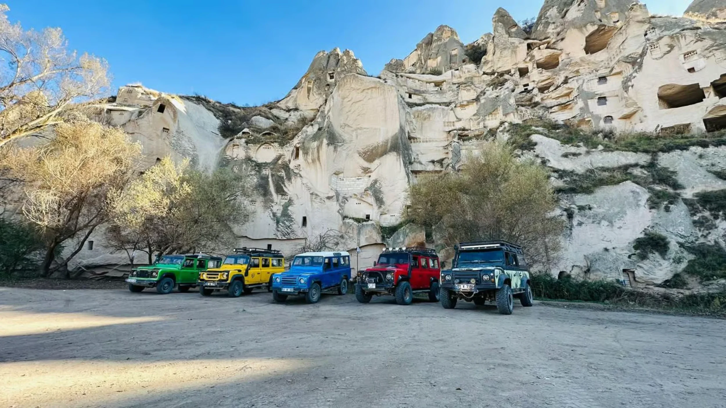 Five colorful off-road vehicles parked in front of a cliffside with numerous cave dwellings carved into the rock, and some trees with bare branches.