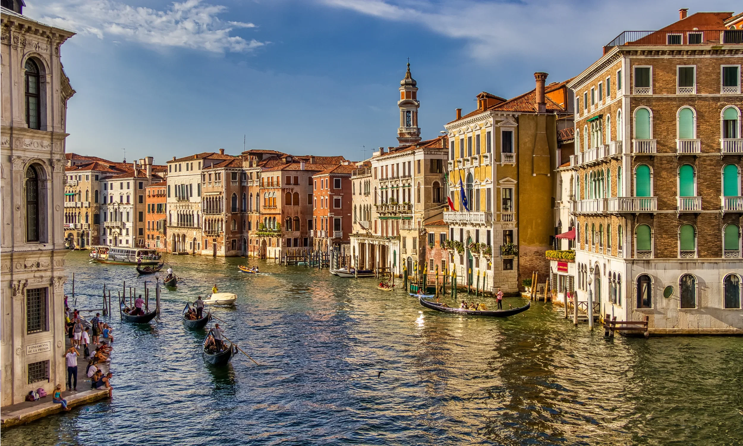 Gondolas on a canal in Venice, Italy, with historic buildings and a church tower in the background under a partly cloudy sky.