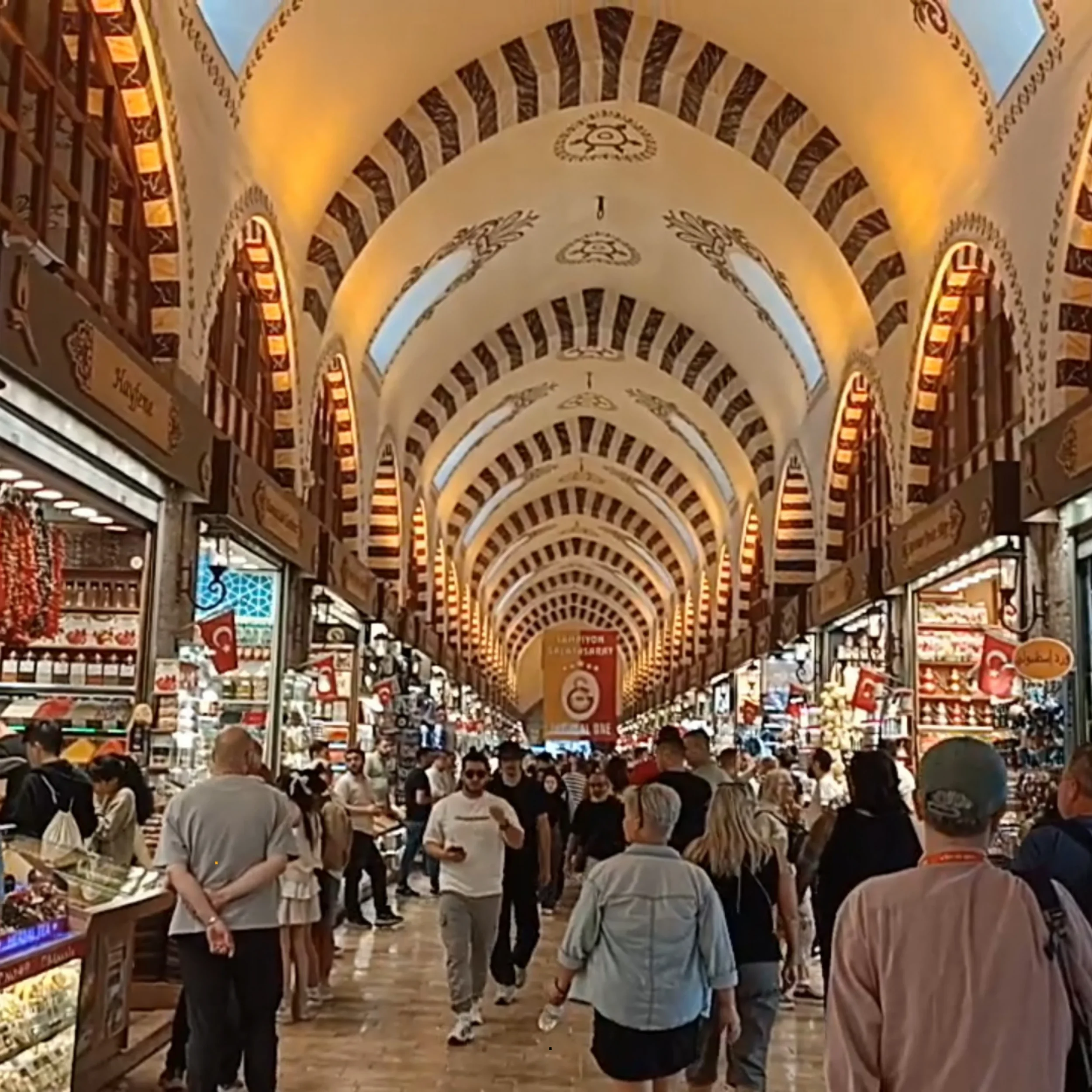 People shopping in a busy indoor market with arches and decorative elements overhead, multiple stalls displaying various goods, and signs in Turkish.