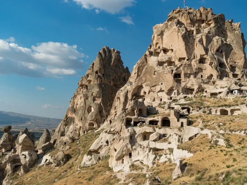 Rock formations with ancient cave dwellings carved into the stone, set against a bright blue sky with a few clouds, in Cappadocia, Turkey.