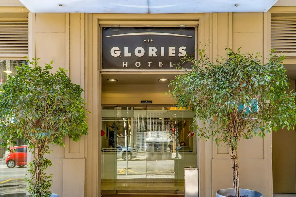 Entrance to Glories Hotel with glass doors, flanked by two potted trees, and a black sign above with white lettering.