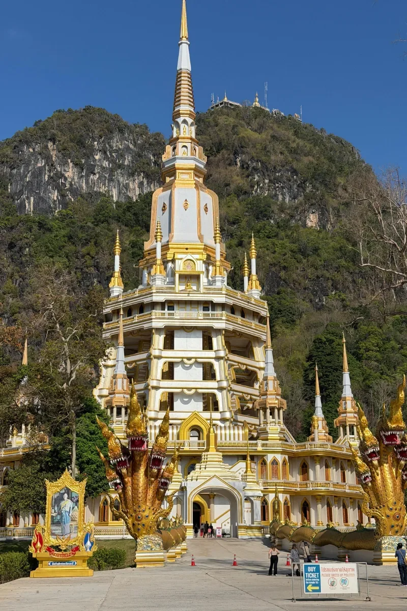A tall ornate temple with a golden spire, featuring multiple layers with gold and white accents, surrounded by mythical serpent statues, set against a hillside with greenery and a blue sky.