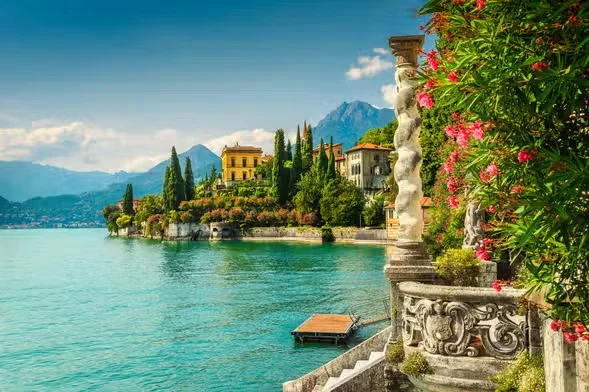 A scenic view of a lakeside town with colorful houses, lush trees, and mountains in the background, along with a stone balustrade with pink flowers in the foreground.