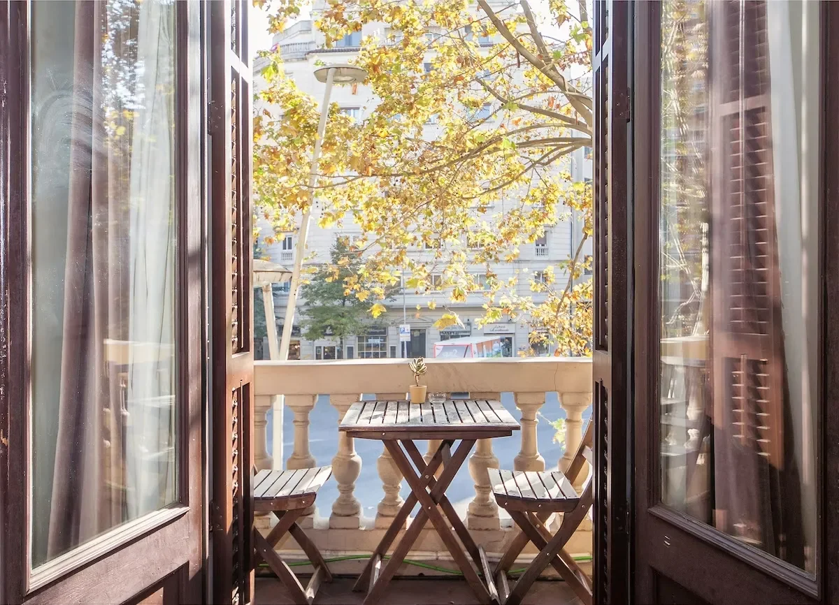 View from a balcony with a small wooden table and two chairs, overlooking a street with trees and buildings, in a city during autumn.