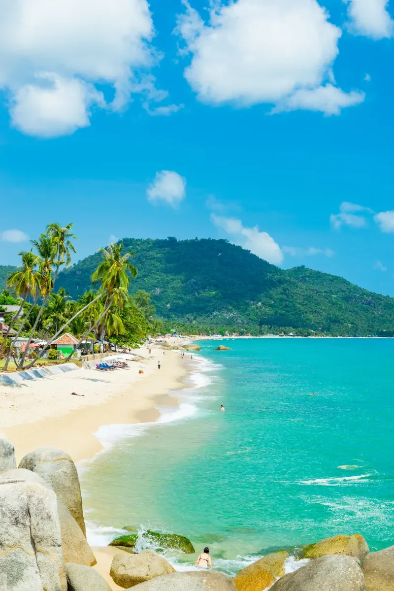 A tropical beach with white sand, turquoise water, palm trees, and a mountain in the background under a partly cloudy sky.